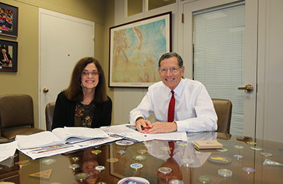 A white woman and a man are sitting at an office table, smiling at the camera.